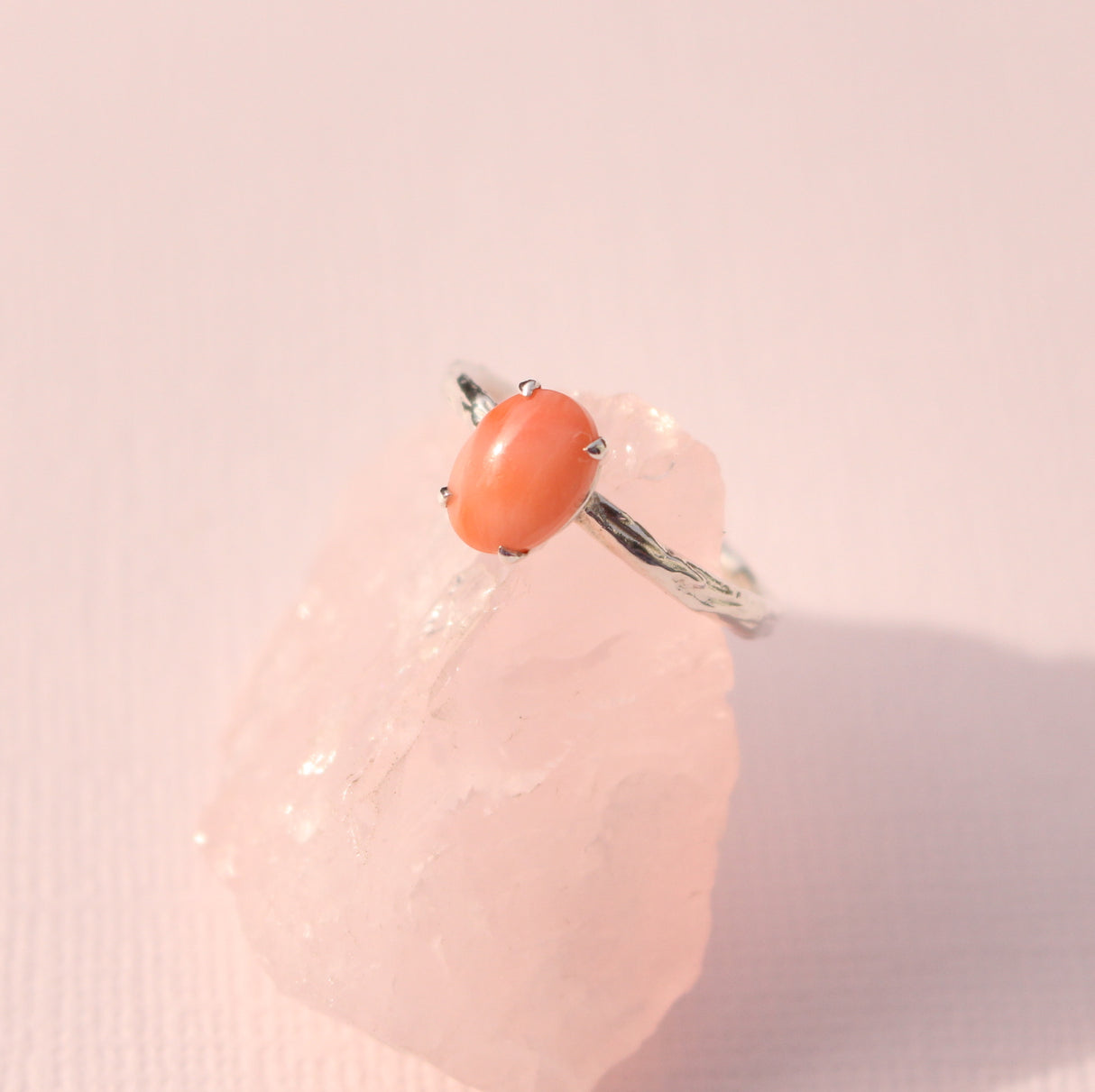 An oval-shaped ring with a coral-colored gemstone in the center, set in a white gold band, displayed on a pink crystal.