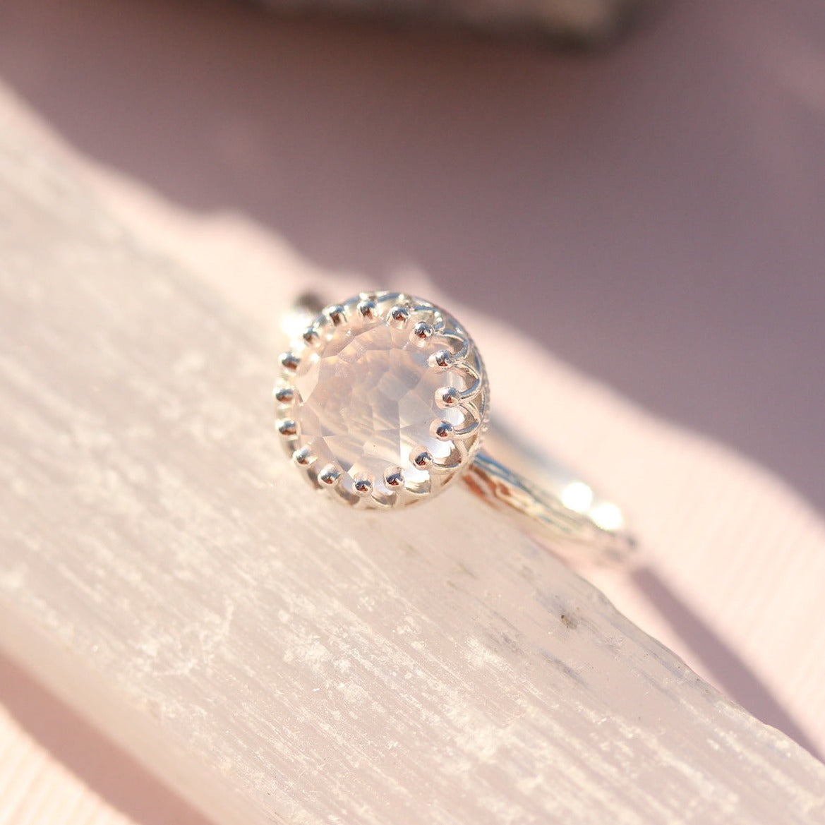 A silver-colored open band ring with a central rose-colored gemstone, displayed on a textured surface.