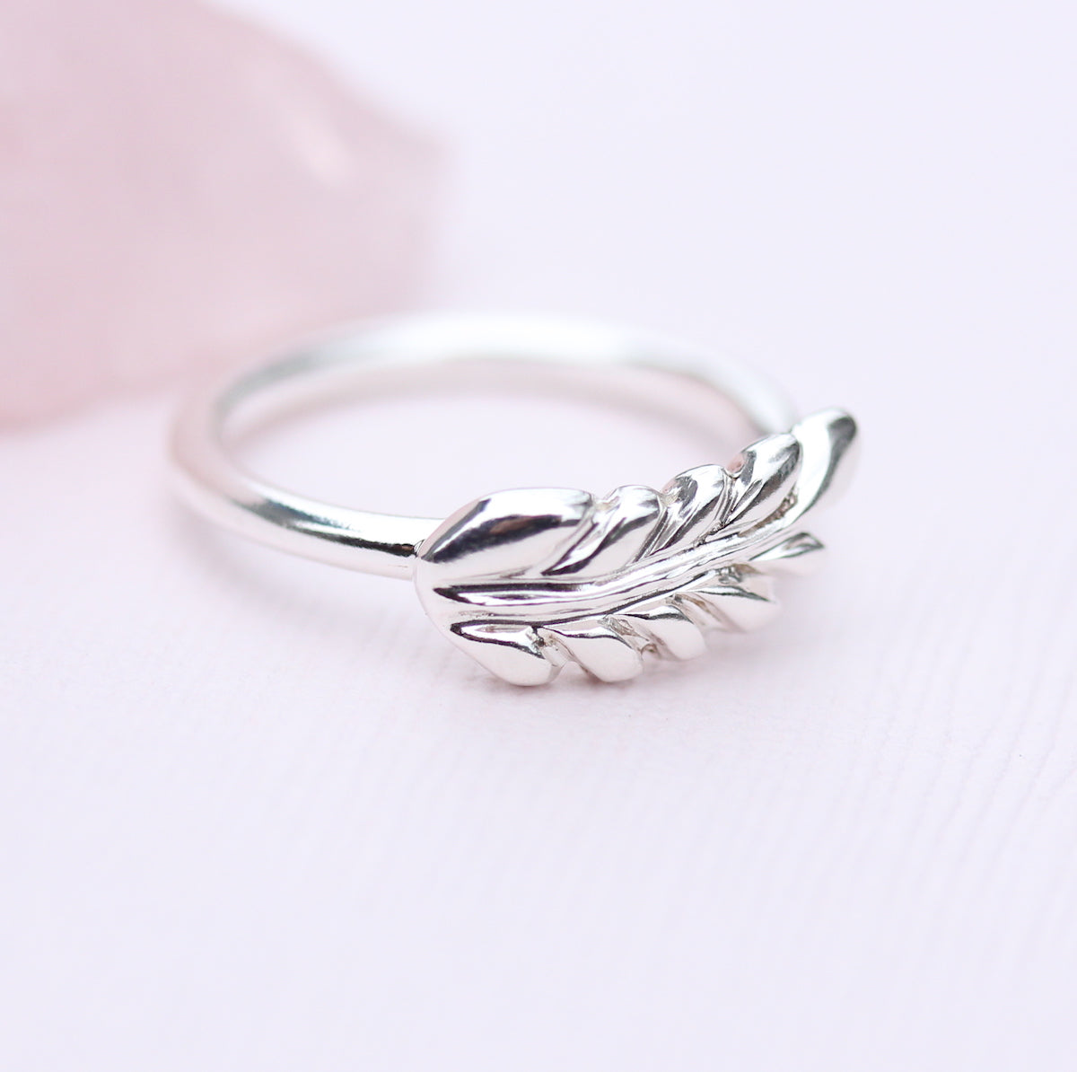 A silver ring with a hand-carved fern motif, set within a metal cutout, on a white background with a pink crystal in the background.