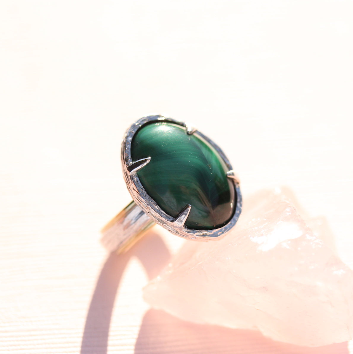 A two-tone ring with a green malachite stone set in a sterling silver band with 18k yellow gold details, displayed on a pink crystal.
