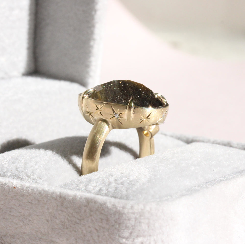 A gold-colored onyx and rutilated quartz ring with small stars on the band, displayed in a ring box.
