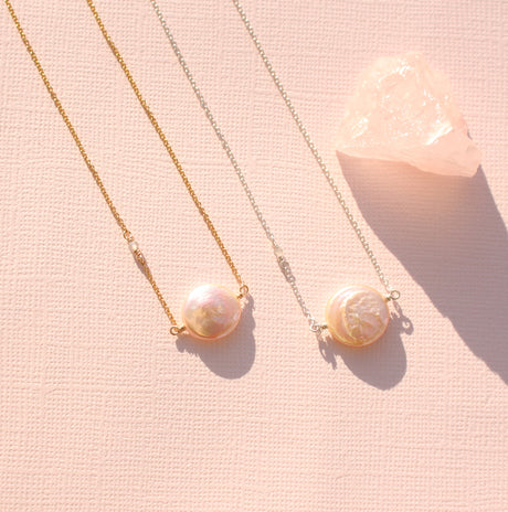 Two pearl necklaces, one with a gold chain and pink pearl, and the other with a silver chain and grey pearl, displayed on a pink background with a crystal to the side.