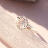 A silver-colored open band ring with a central rose-colored gemstone, displayed on a textured surface.